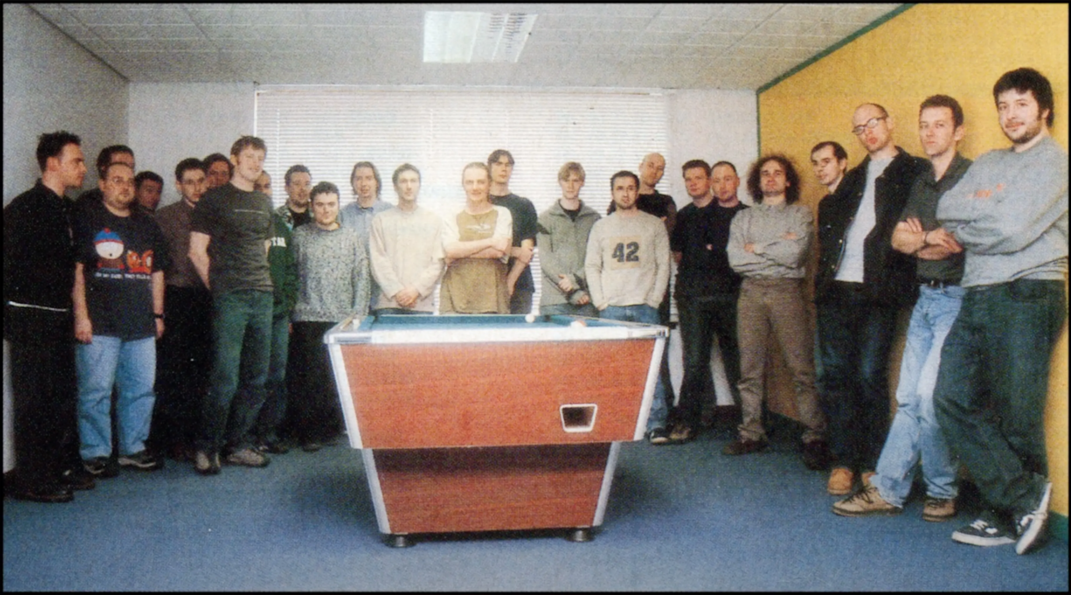 A period photo of the GTA III development team, around twenty people standing around a pool table in the office.