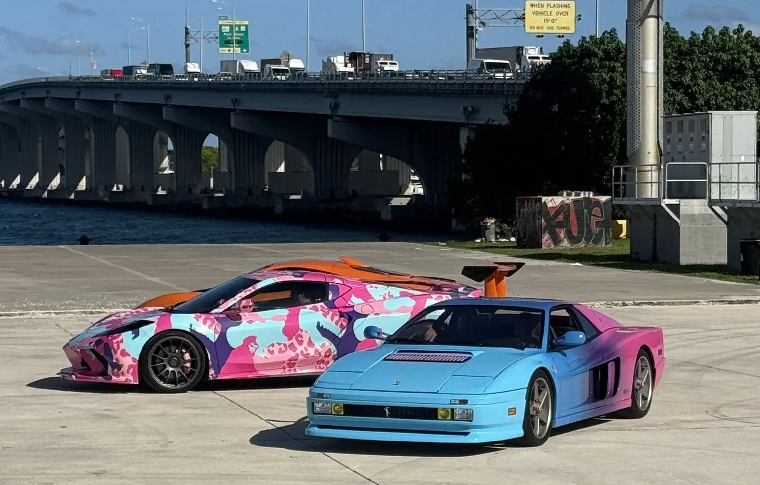 Dos deportivos decorados con la estética de GTA VI aparcados junto a un puente de Miami: uno con manchas rosas, azules y moradas y otro azul degradado a rosa.