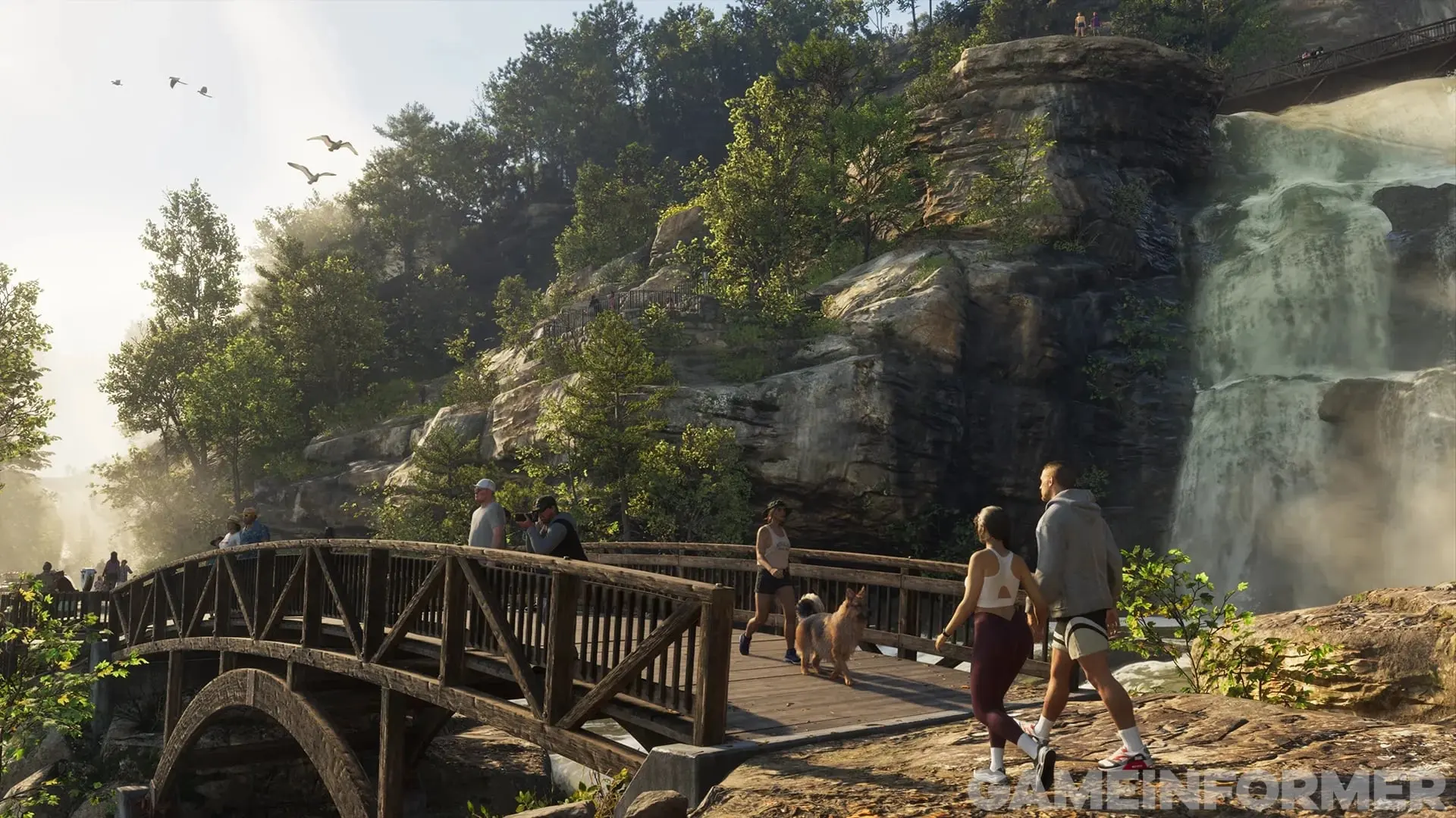A wooden bridge beside a waterfall in a wooded area, with a group of hikers crossing and birds in the air.