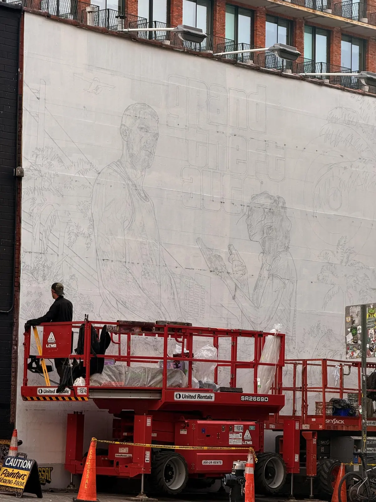 A white Brooklyn building wall with the pencil outline of the GTA VI mural: Jason, Lucia holding a pistol, palm trees and the Grand Theft Auto logo, with red scissor lifts in front.