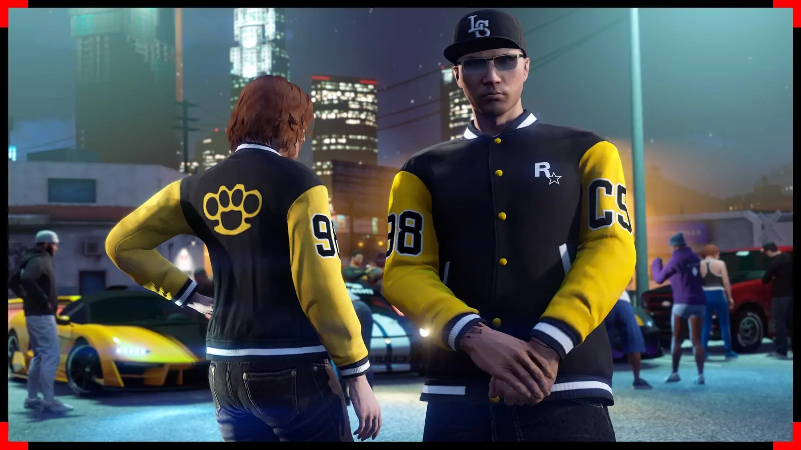 Dos personajes de GTA Online con la chaqueta universitaria Rockstar Community Series en una reunión de coches nocturna.
